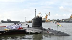 A Kalvari-class Indian Navy submarine manoeuvres into harbour with crew members on deck, against the backdrop of a busy port. (Photo: PTI)