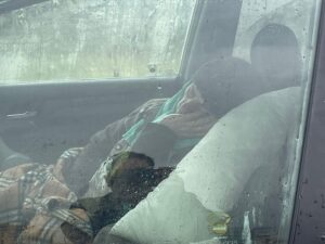 Inside a parked car, an elderly rests against a pillow, face turned away from the rain-streaked glass.