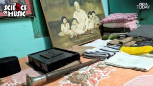 A shehnai rests in the foreground at Ustad Bismillah Khan's Varanasi home, beside a harmonium and a stack of folded clothes. The painting in the background shows the maestro seated with three children, a quiet glimpse into the life he lived between his concerts at the Red Fort and the Cannes Art Festival. (Photo: PTI)